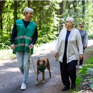 Vanhempi nainen juttelee Tukikoirakko-tunnusta kantavan naisen kanssa, joka taluttaa koiraa.