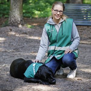 Tukikoirakko-liivi kaulassa oleva koira lemää maassa. Kyykyssä sitä silittää hymyilevä nainen Tukikoirakko-huomioliivissä.